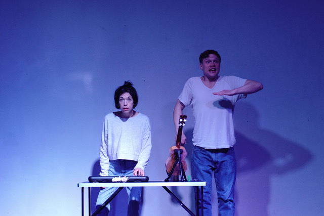 Schauspieler:innen Verena Stier und Justus Verdenhalven vor einer bläulich angeleuchteten Wand, vor Ihnen ein Keyboard und eine Violine