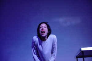 Schauspielerin Verena Stier mit aufgerissenem Mund vor blau beleuchteter Wand